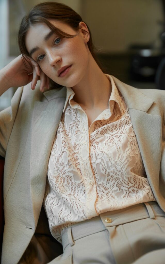 A natural-looking young woman wearing a delicate lace top layered under a neutral-toned suit. Theme Lace Tops and Outfits