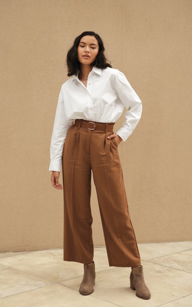 A natural-looking young woman wearing brown pants styled with a belt and ankle boots for a flattering, put-together look. Theme brown pants outfit for women