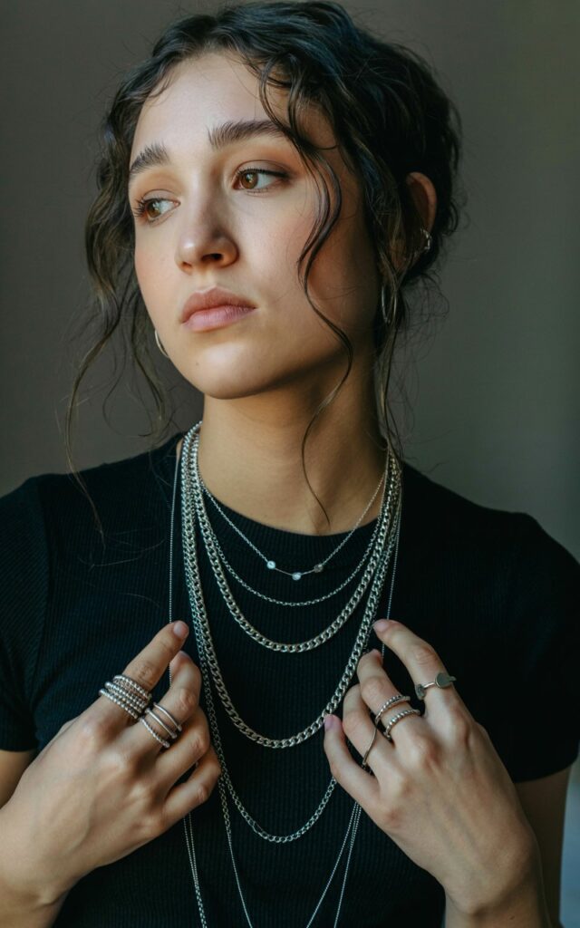 A natural-looking young woman wearing chunky chain necklaces and multiple silver rings as statement accessories. Theme emo outfits