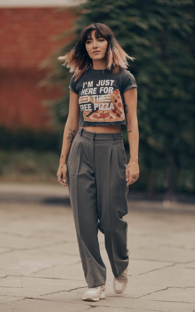 A natural-looking young woman wearing tailored trousers styled with a cropped graphic tee and sneakers. Theme tomboy outfits
