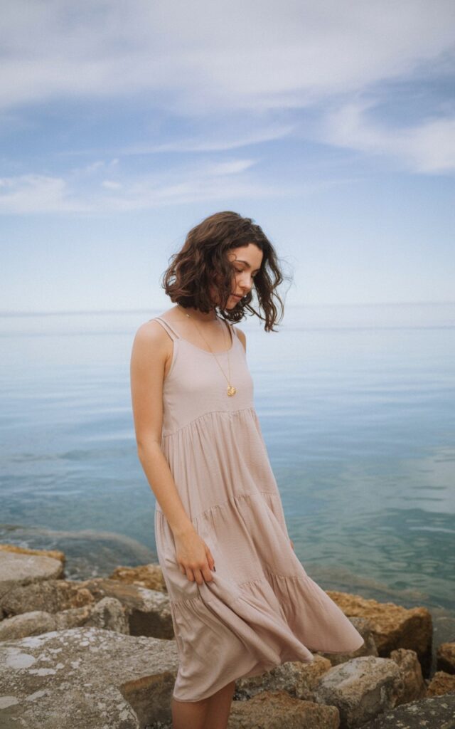 A photo of a natural-looking young woman wearing a tiered midi dress that flows in the wind. She has wavy, dark hair and wears a pendant necklace. The woman stands on a rocky beach with a backdrop of calm water and a distant horizon. The sky has a few clouds. The lighting is soft.
