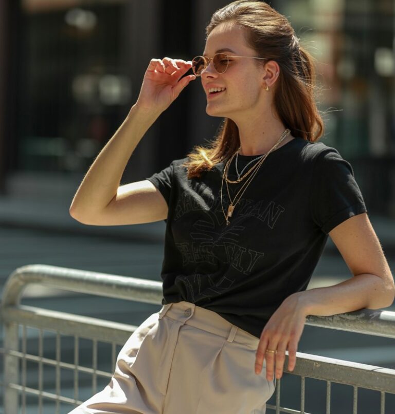 Full-body of a white-skinned model wearing a black graphic tee tucked into beige tailored shorts, white sneakers, layered necklaces. She leans on a city railing, adjusting her sunglasses, smiling softly. Soft sunlight, realistic editorial style, candid natural pose.