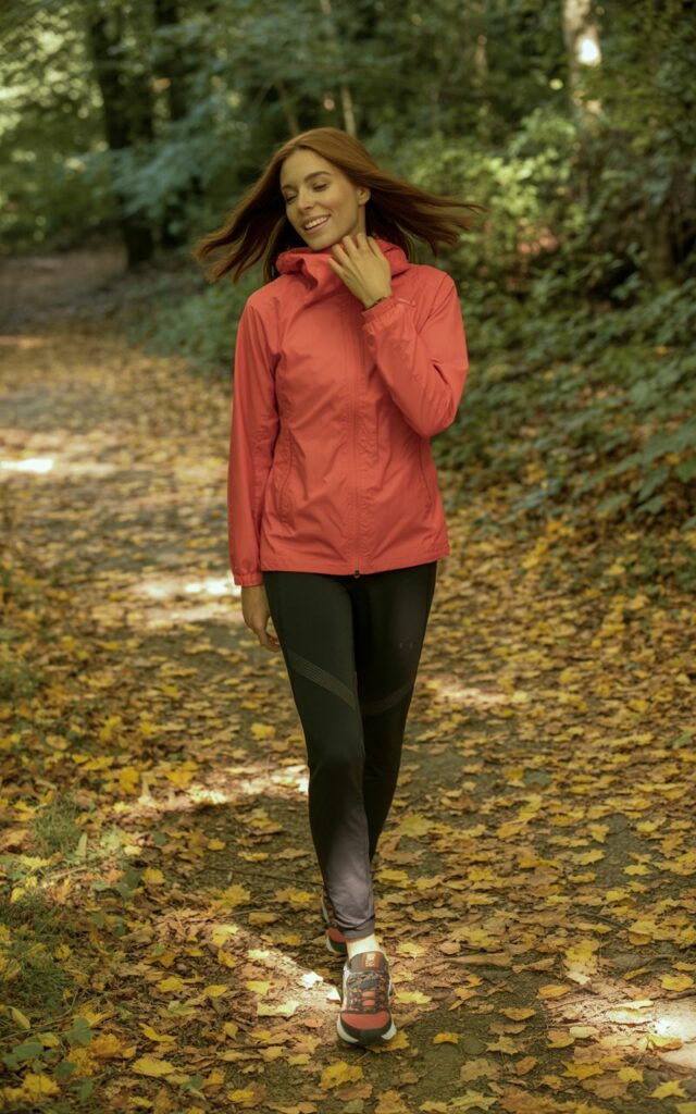 Full-body realistic shot of a white-skinned female model with symmetrical features wearing a bright windbreaker, black leggings, and trail sneakers. Standing mid-hike on a leafy forest path, adjusting her hood with a soft smile. Sunlight filtering through the trees, casting natural shadows. Realistic hair highlights blowing slightly in the breeze. Shot on a 50mm lens, editorial candid style, soft depth of field, natural color grading.