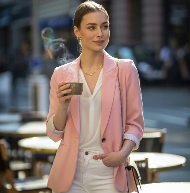Full-body realistic white-skinned female model with symmetrical face, soft makeup, and healthy fit body. Wearing a pastel pink tailored blazer over a white blouse, white skinny jeans, and nude heels, accessorized with a delicate gold necklace and a small crossbody bag. Outdoor city café setting with tables, chairs, and a sunny morning vibe. Natural sunlight highlighting textures and shadows. Model standing casually, holding coffee, smiling softly. Candid editorial photography, 50mm lens, soft depth of field.