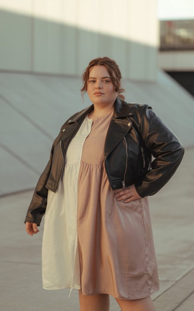 "A natural-looking young curvy woman wearing a cropped moto jacket over a flowy dress. Theme: curvy outfits."