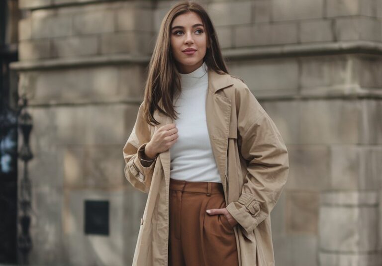 A natural-looking young woman wearing brown pants styled with a lightweight trench coat for a timeless, classic outfit. Theme: brown pants outfit for women