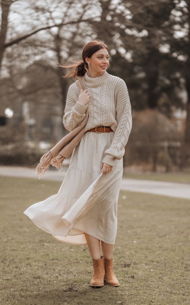 A natural-looking young woman wearing a belted sweater layered over a white skirt with ankle boots. Theme white skirt outfit winter