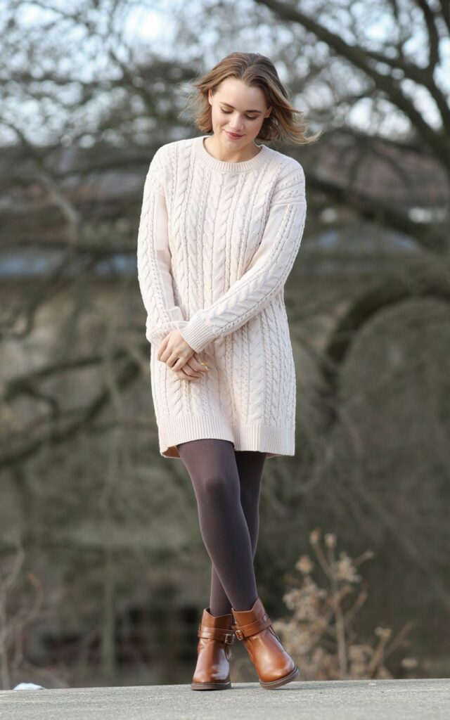 A natural-looking young woman wearing a cozy sweater dress paired with tights and casual ankle boots. Theme cute casual winter outfits