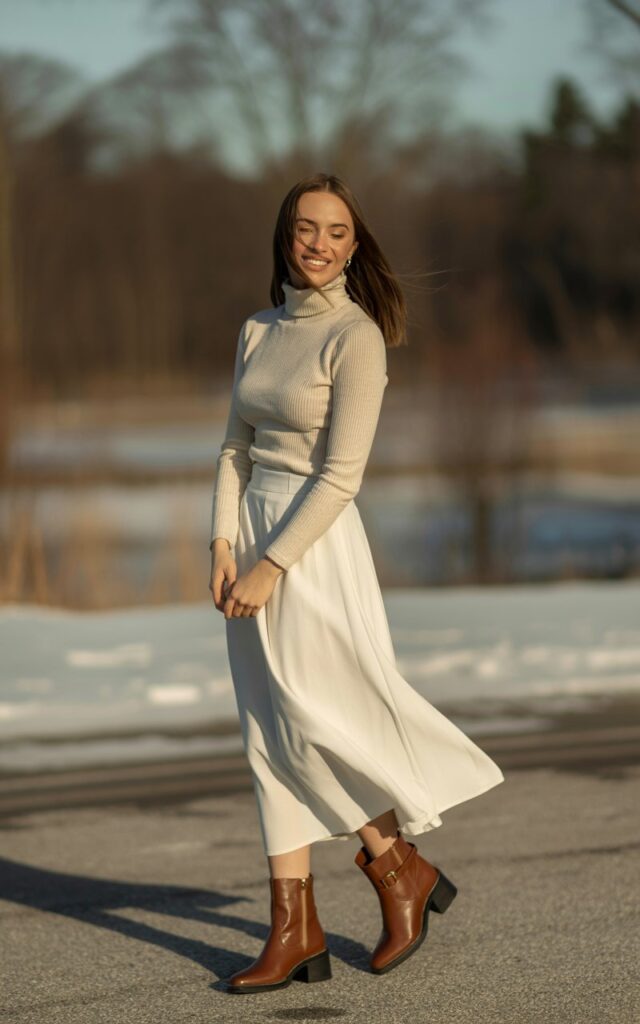 A natural-looking young woman wearing a fitted turtleneck tucked into a flowy white skirt with winter boots. Theme white skirt outfit winter
