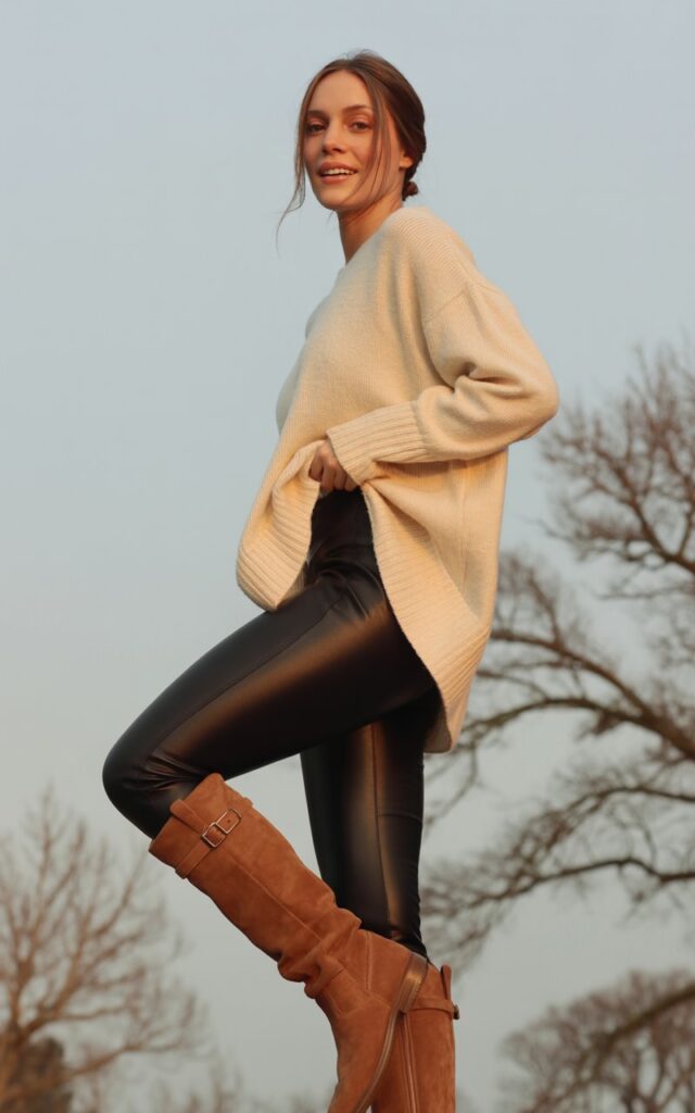 A natural-looking young woman wearing black leggings paired with a long oversized sweater and knee-high boots. Theme legging outfits winter