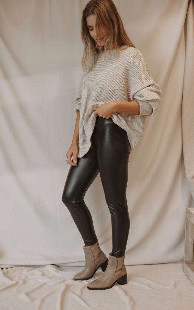 A natural-looking young woman wearing faux leather leggings paired with a chunky knit pullover and neutral boots. Theme legging outfits winter