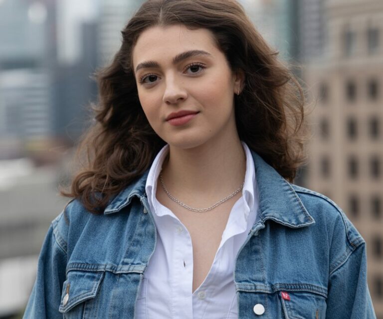 A photo of a young woman with natural-looking features wearing a denim jacket and matching denim jeans. Her dark hair is styled in loose waves. She wears a white shirt underneath the jacket and has a necklace. The background is a blurred cityscape with buildings. The lighting is soft.