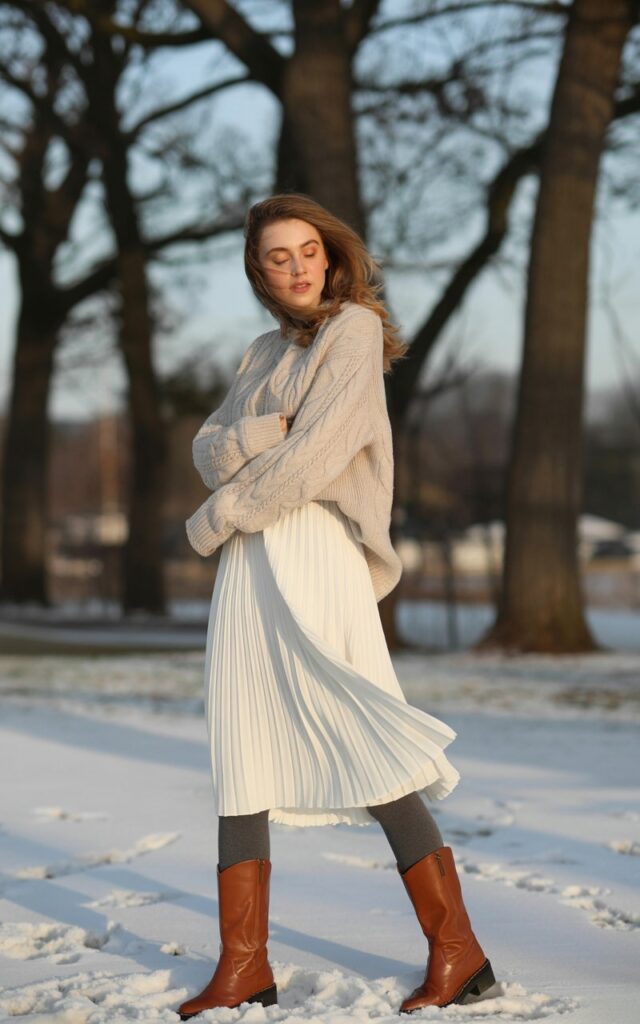 A photograph of a young woman wearing a cozy winter outfit featuring a white pleated midi skirt as the centerpiece. She pairs the skirt with warm charcoal gray tights, knee-high leather boots in rich brown, and a cream cable-knit sweater with an oversized, dress-like silhouette that creates elegant layers. Her natural makeup and softly tousled hair complement the effortless styling, while she stands against a backdrop of bare winter trees with patches of snow on the ground. The lighting is soft and diffused, creating a gentle winter atmosphere that highlights the texture contrasts between the flowing white fabric, knitted wool, and smooth leather.