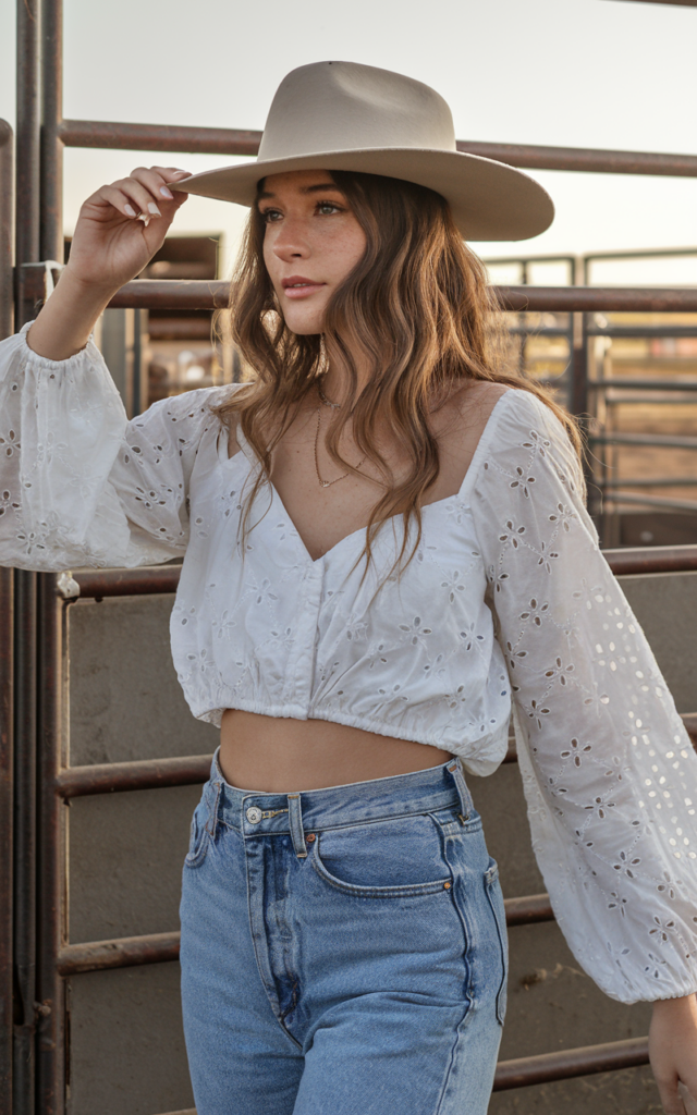 A natural-looking young beautiful woman wearing a breezy white eyelet blouse tucked into high-rise flare jeans, a wide-brim felt hat, and stacked-heel cowboy boots, styled with soft waves for a golden hour rodeo setting.